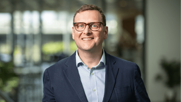 A man wearing a suit and glasses smiles at the camera. He's in a corporate environment.