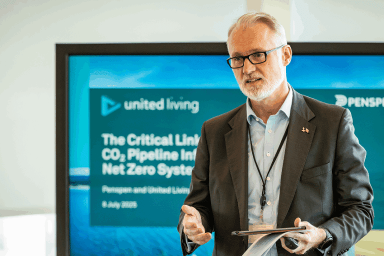 man in suit with grey hair and glasses stands in front of presentation screen