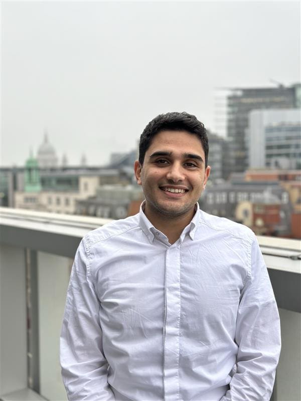 A young man in a shirt smiling in front of the London skyline.