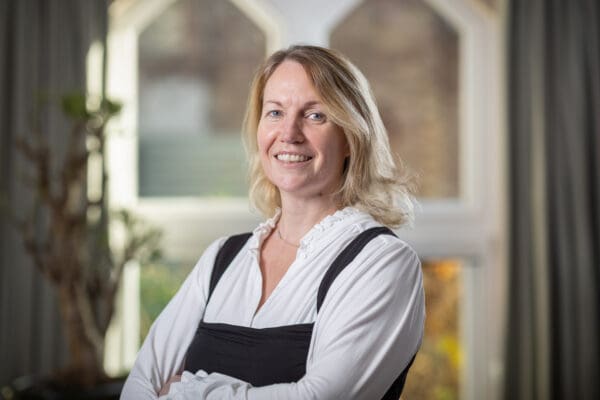 A woman with blonde hair and wearing a white blouse stands in front of a window. She's smiling and has her arms crossed on front of her body.