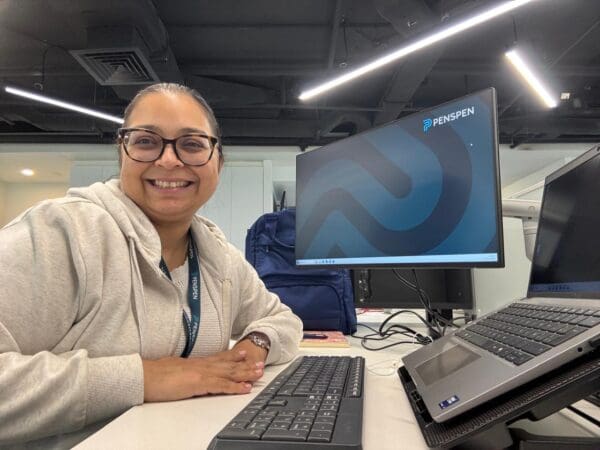 An image of a woman sitting at her desk in front of her computer. She's smiling and has the Penspen logo on her screen.
