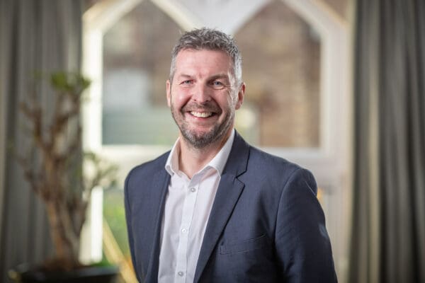 A main wearing a suit standing front of a window. He is smiling.