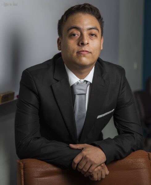 An image of a young man wearing a suit. He is leaning forward onto a desk with his hands crossed.