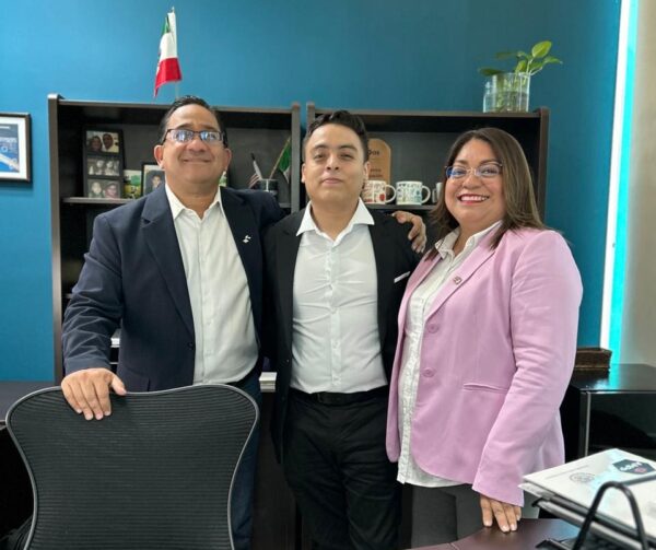 Two men and a women stand behind a desk in an office. They are dressed smartly in suits.