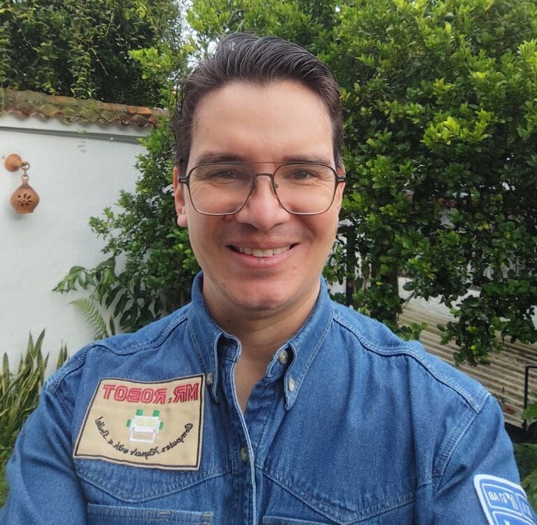 An image of a man in his thirties with dark hair and glasses. He is smiling at the camera and wearing a casual blue shirt