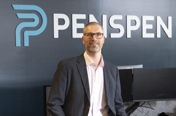 A white male wearing glasses and a suit sits against a desk, and smiles at the camera. In the background is a large Penspen sign on the wall.