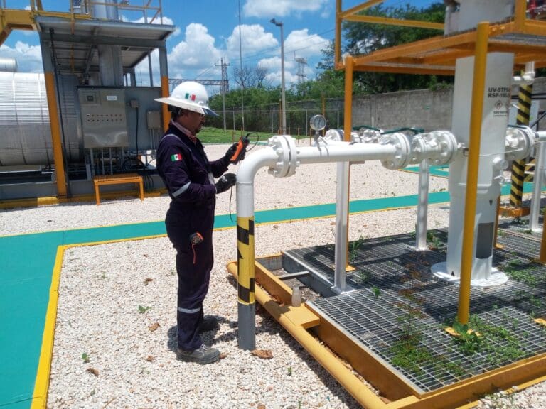 Technician in protective equipment uses a handheld monitoring device to inspect industrial piping at an outdoor gas processing facility.