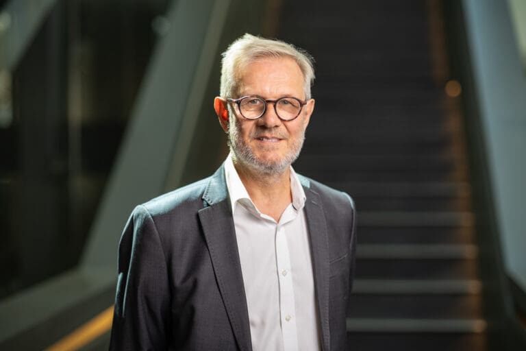 Am image of a man wearing glasses and a grey suit, with no tie. He stands in front of a staircase.